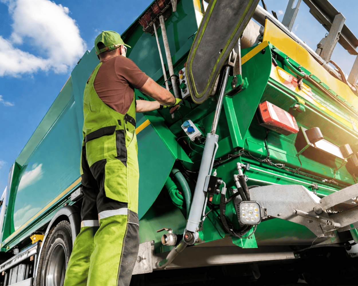 Sanitation worker operating the rear of a green garbage truck during waste collection.