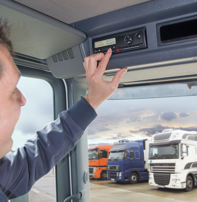 Driver adjusting a digital tachograph inside a truck cabin, with parked trucks visible outside through the window.