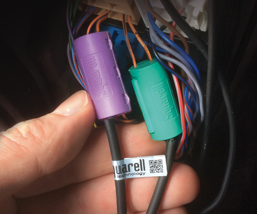 Close-up of a hand holding two colored Sumitomo electrical connectors (purple and green) attached to wiring inside a vehicle.