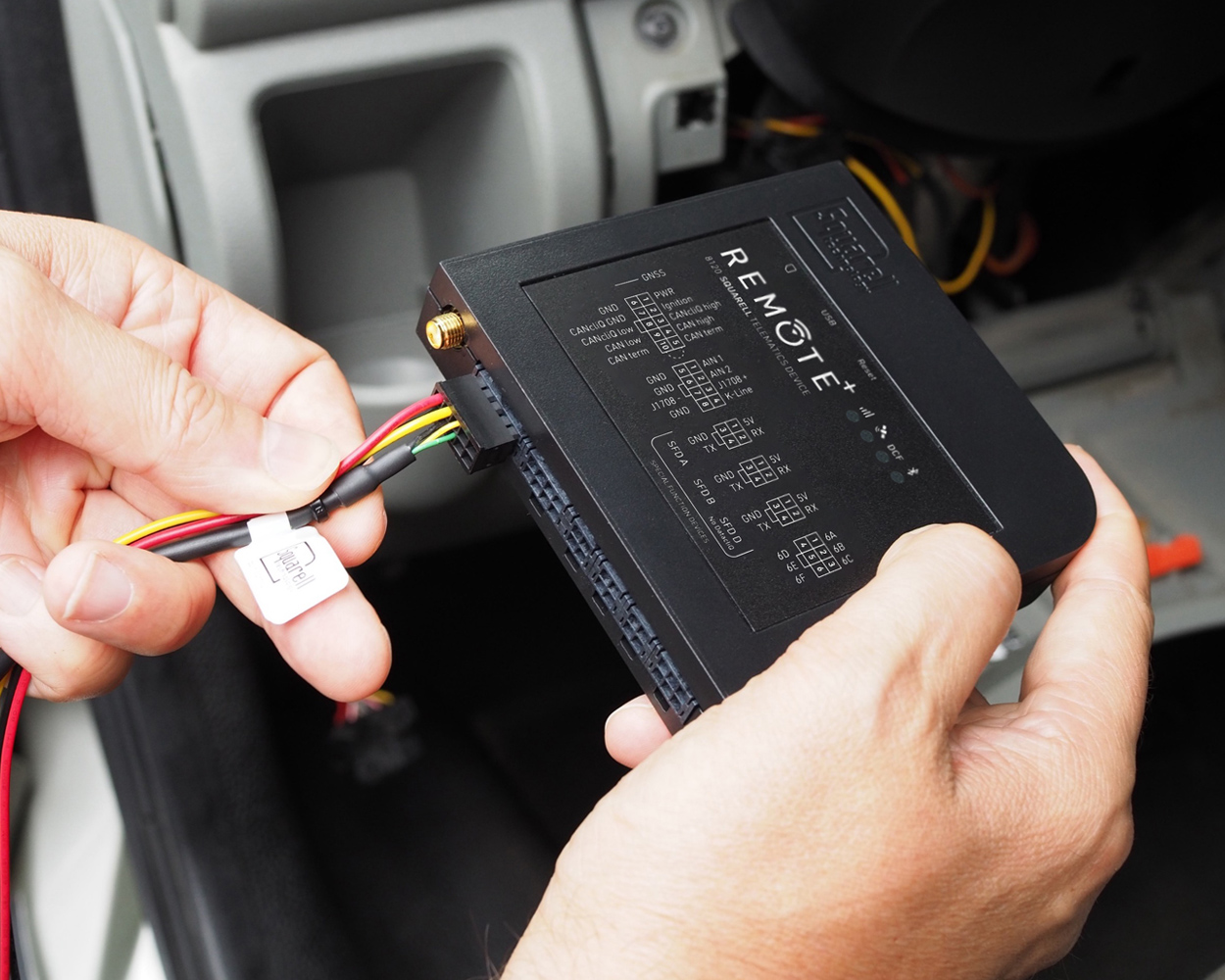 Hands connecting wiring to a REMOTE+ vehicle tracking device inside a car dashboard.