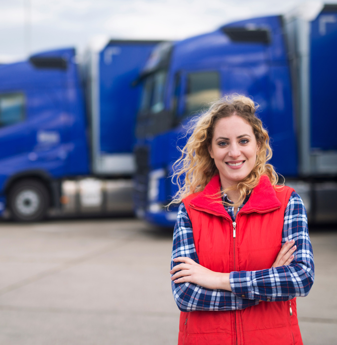 Smiling woman standing with arms crossed in front of blue trucks in a logistics yard.