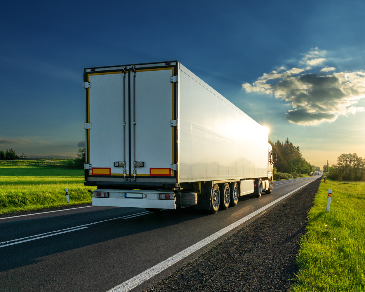 White cargo truck driving on a rural road through green countryside at sunset.