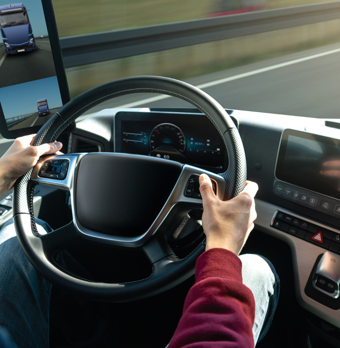 Driver operating a modern truck with a digital dashboard and side camera display while driving on a highway.