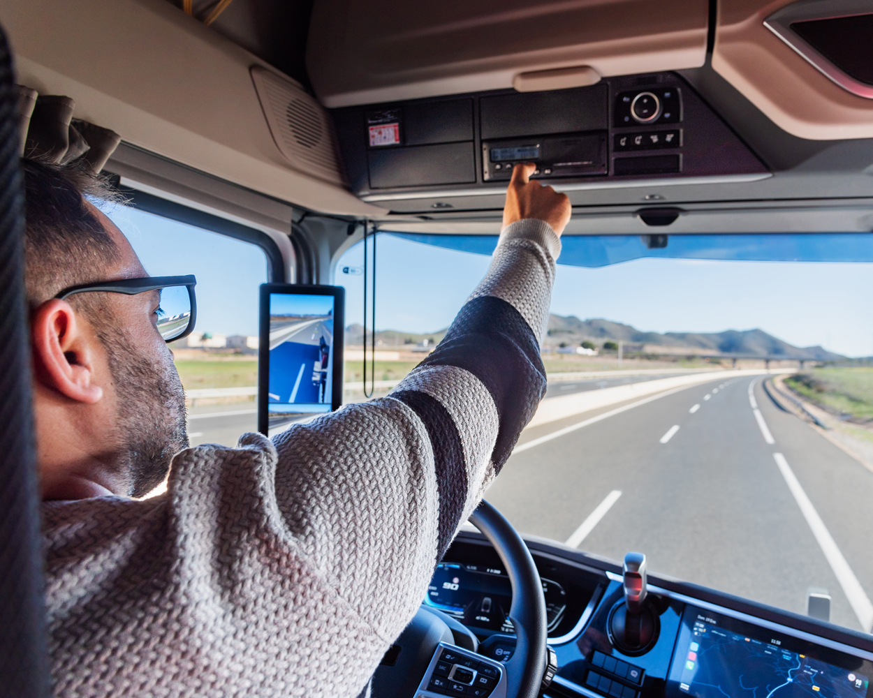 Truck Driver Adjusting Controls While Driving Truck driver reaching toward the overhead control panel while driving on a highway through an open landscape.