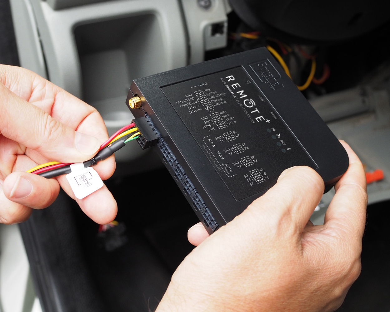 REMOTE+ Telematics Device Installation A technician connects wiring to a REMOTE+ telematics device inside a vehicle, preparing the unit for installation and data communication.