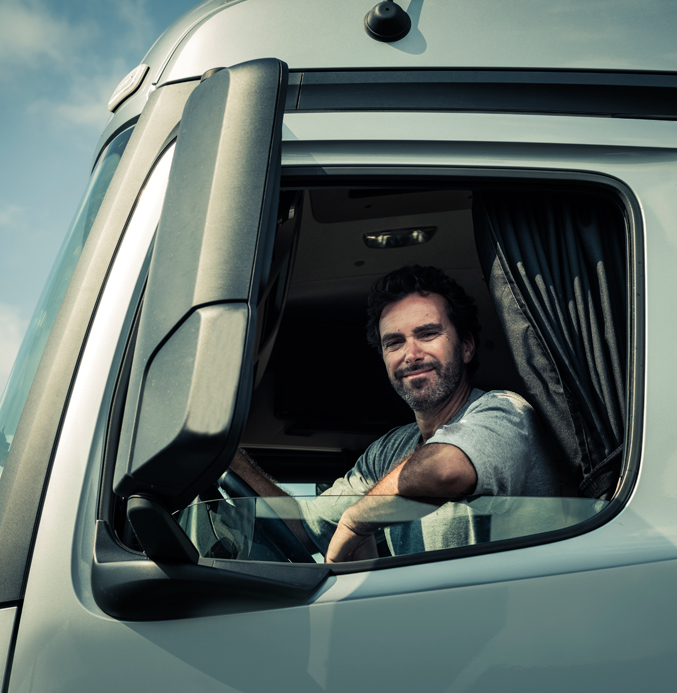 Professional Truck Driver in a Modern Semi-Truck Smiling truck driver sitting in the cab of a modern semi-truck, looking out of the open side window during daylight.