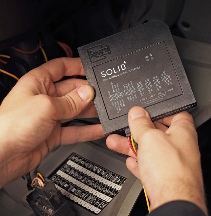 SOLID+ Vehicle Telematics Device Installation A technician installs a SOLID+ telematics device inside a vehicle, holding the compact black unit near the vehicle’s electrical connection panel and wiring harness during setup.