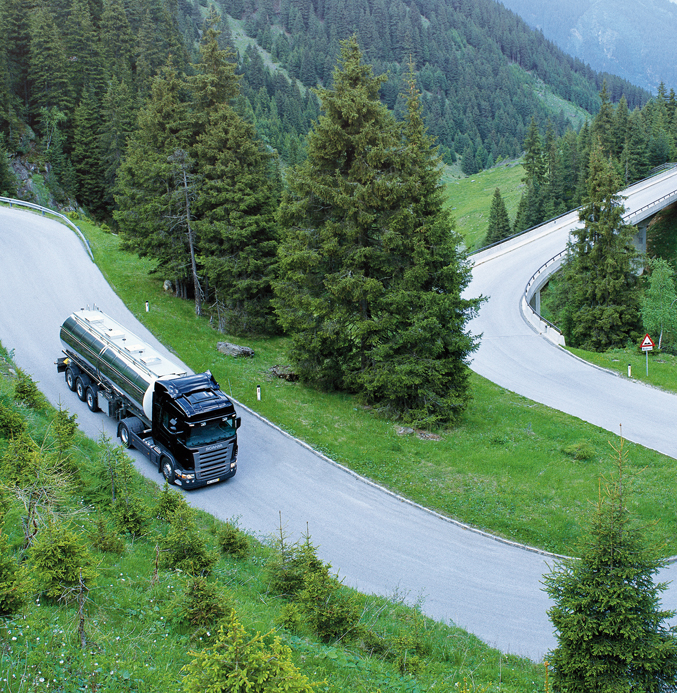 Tanker Truck Navigating a Scenic Mountain Road Black tanker truck driving on a winding mountain road surrounded by dense pine forests.