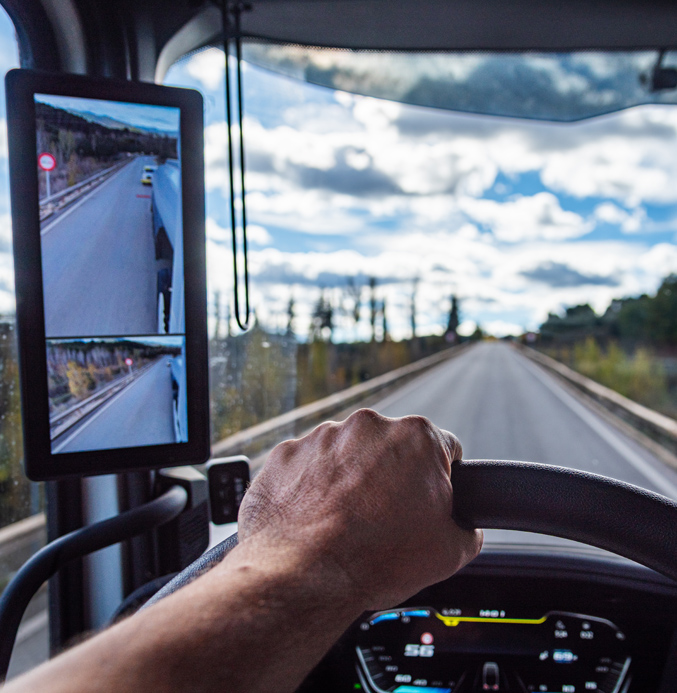 Truck Driver Using Digital Side Mirrors on Open Road Driver’s perspective inside a truck with a hand on the steering wheel, looking down a straight road with a digital side mirror display.