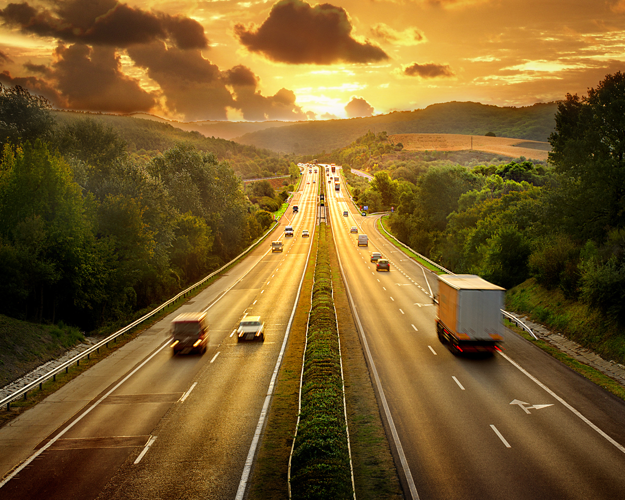 A busy divided highway running through a green countryside at sunset, with cars and trucks traveling beneath a dramatic golden sky.